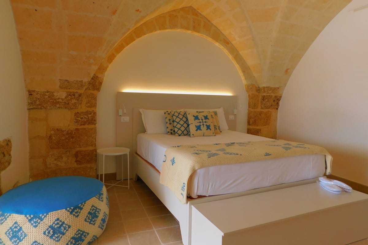A bedroom with a stone wall, a bed with patterned bedding, a bedside table, and a small round chair.