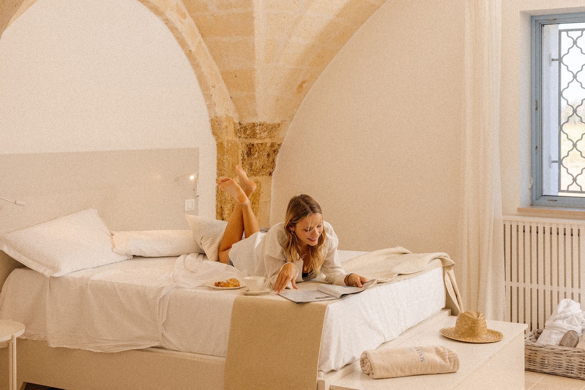 A room with a bed under an arched ceiling, a window with a lattice design, and a basket of items on the floor.