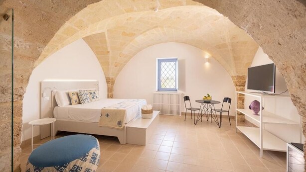 A cozy bedroom with a stone arch, a bed, a small table, a TV, and a window. - Masseria Malvindi (Mesagne)