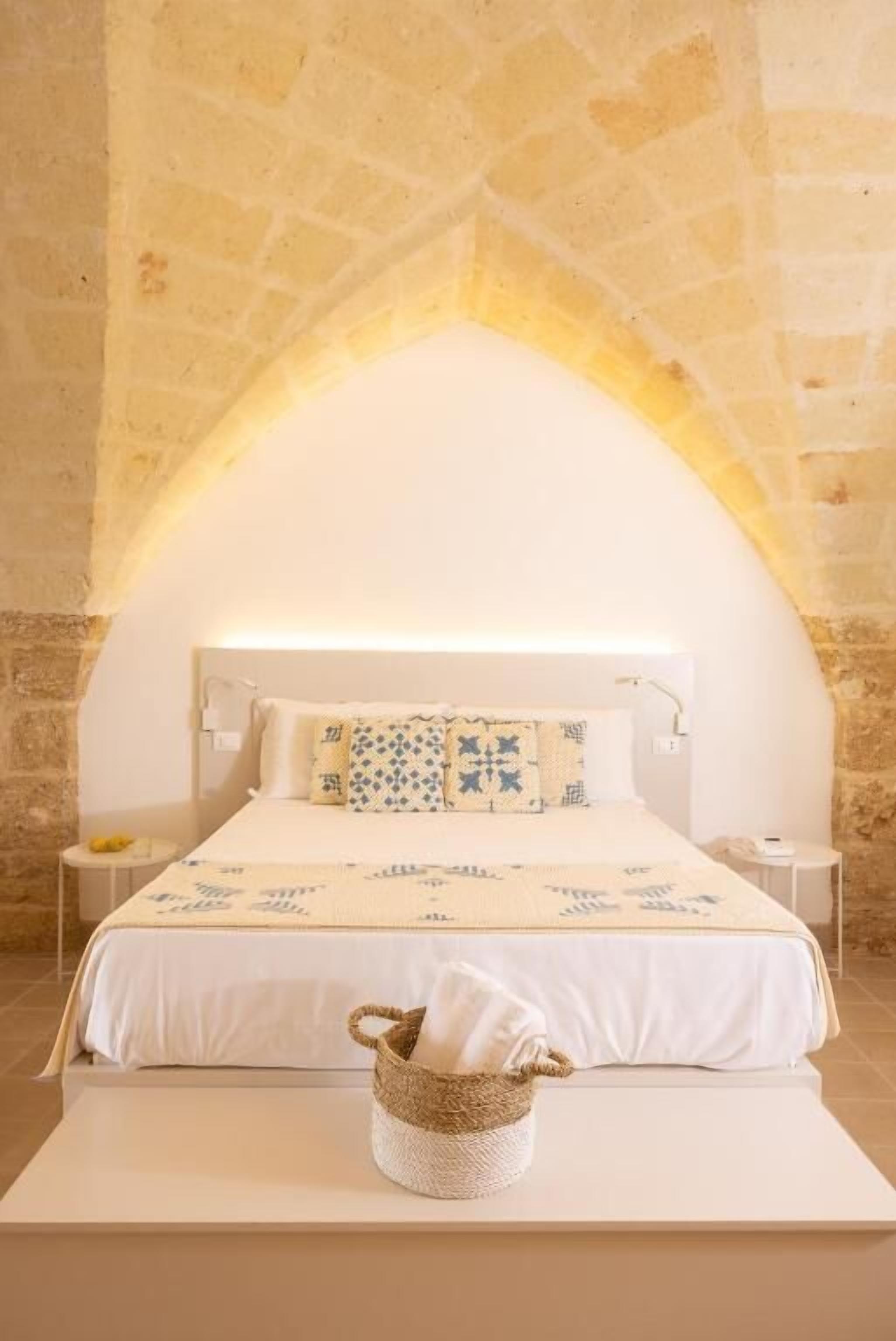 Una cama con cabecera blanca, una colcha estampada y una mesita de noche con lámpara.