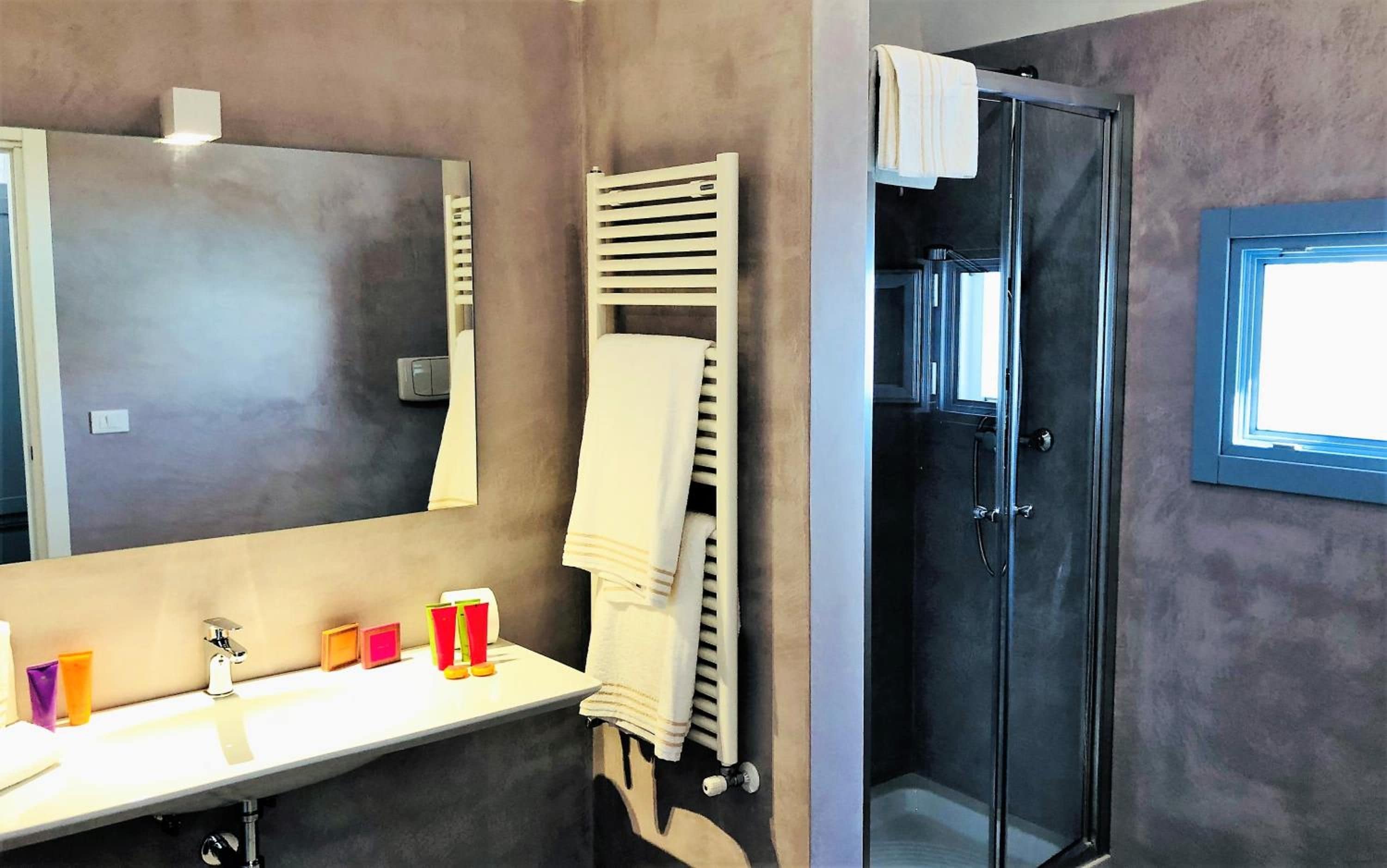 A bathroom with a large mirror, a white sink, a towel rack, a shower enclosure, and a window.