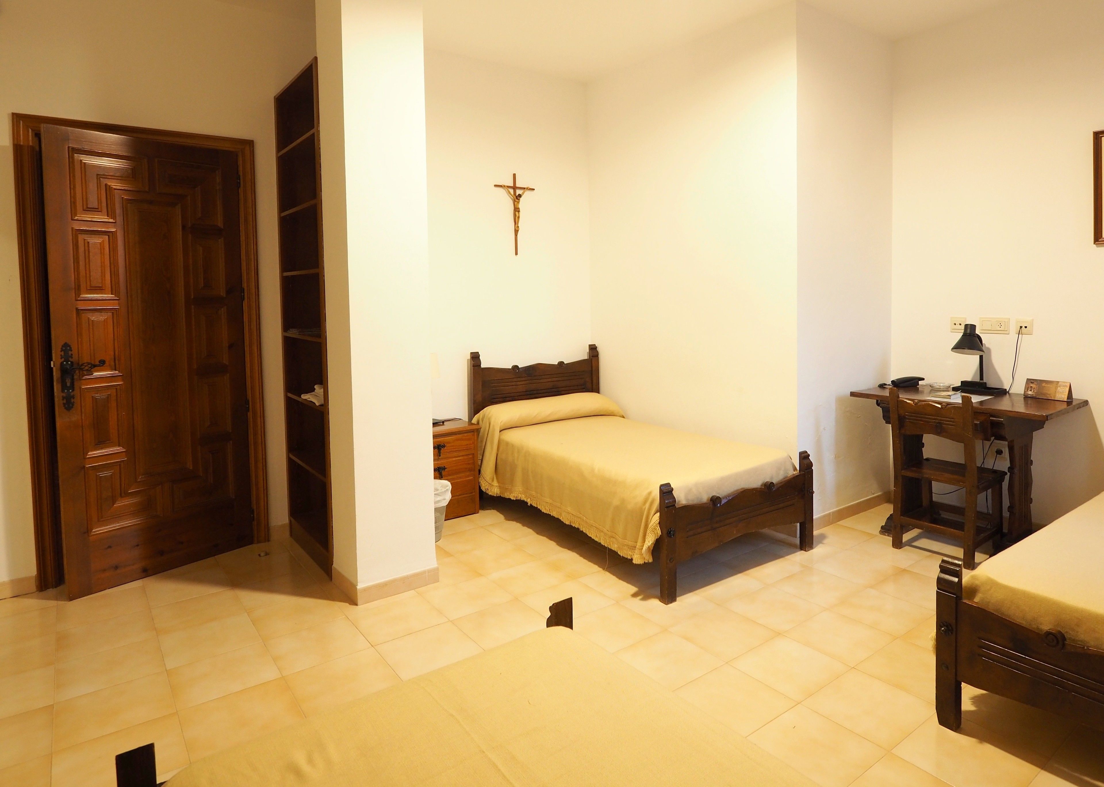 Une chambre à coucher avec un lit en bois, une table de chevet, un bureau avec une chaise, une armoire en bois et un crucifix accroché au mur.