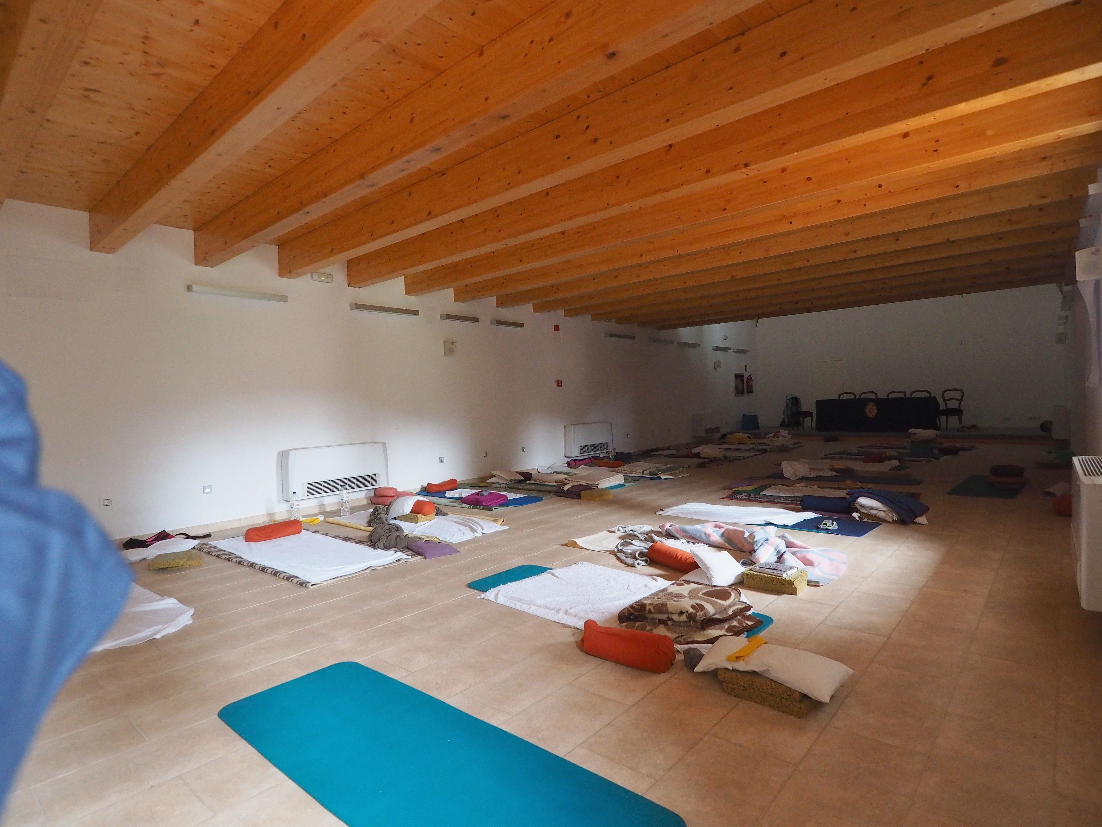Um quarto espaçoso com vigas de madeira no teto, vários tapetes de yoga e alguns almofadas e mantas espalhados pelo chão.