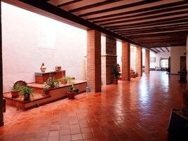 Un corredor espacioso con suelo de baldosas, paredes de ladrillo y vigas de madera en el techo. Hay plantas en macetas a lo largo de las paredes y un banco al fondo.