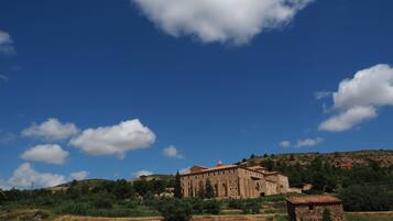 A historic building with a red-tiled roof, surrounded by a field and trees, under a clear blue sky.