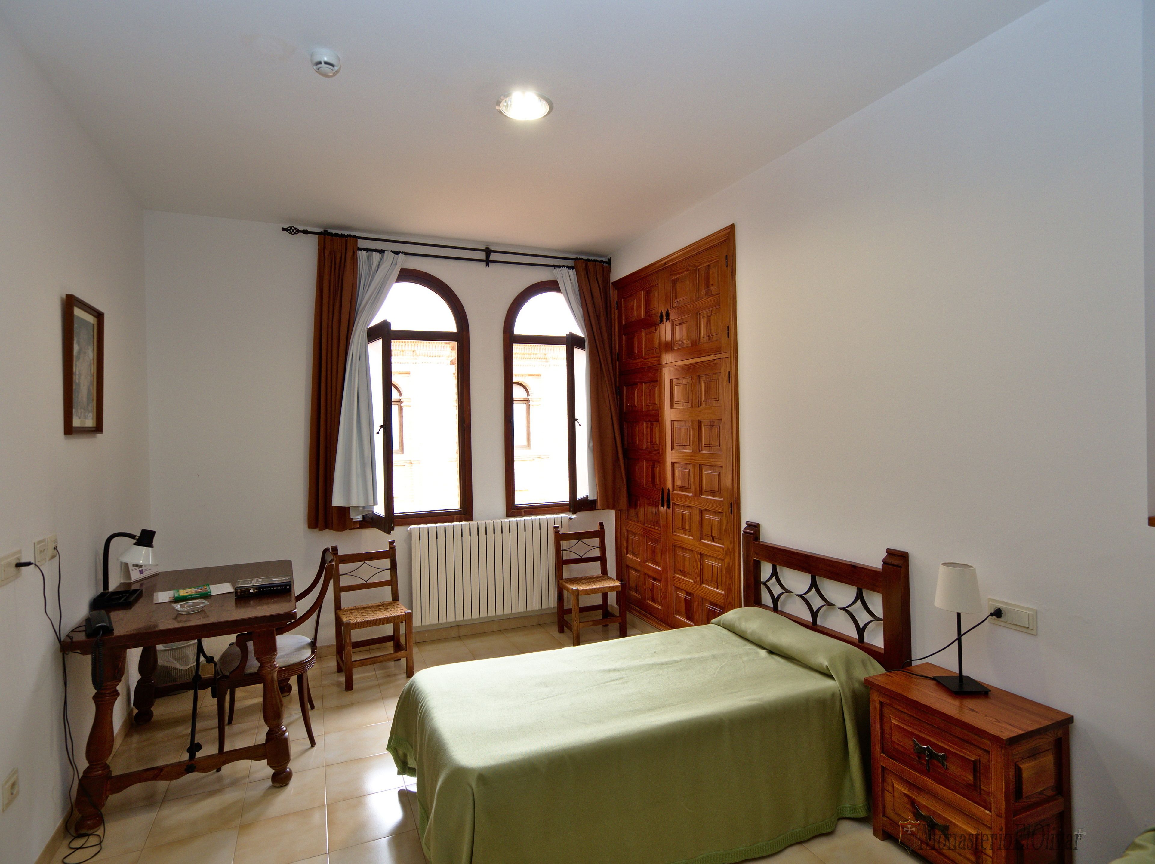 A bedroom with a bed, a desk, two chairs, a radiator, and a wooden wardrobe.
