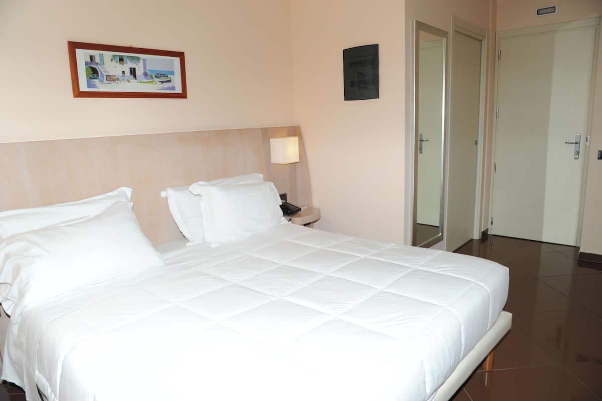 Een hotelkamer met een groot bed, een nachtkastje met een lamp, een ingelijst schilderij aan de muur en een deur naar een andere kamer.