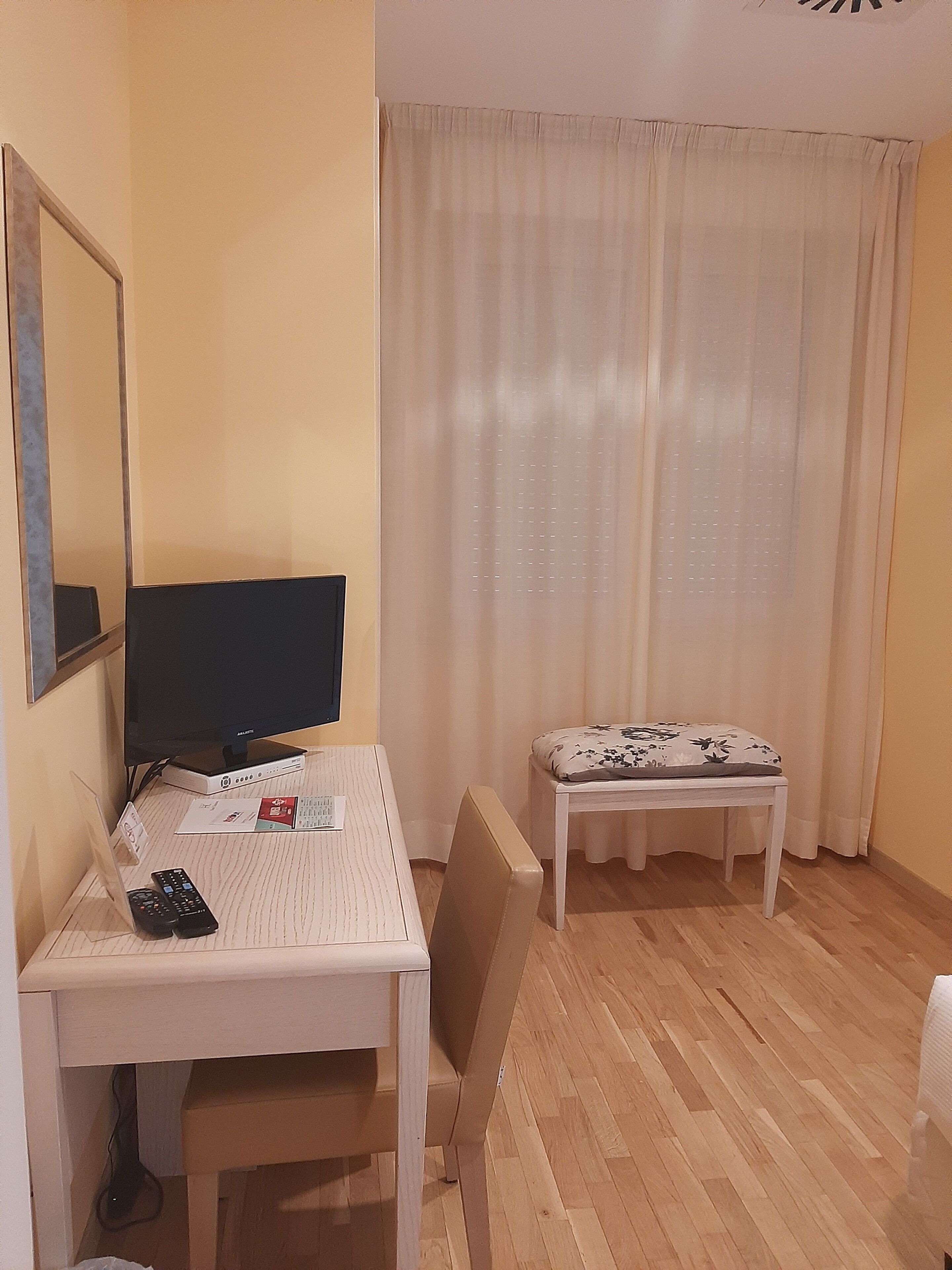 Une pièce comprenant un bureau, une chaise et une petite table recouverte d’une nappe à motifs floraux. Il y a un téléviseur sur le bureau et un miroir au mur.