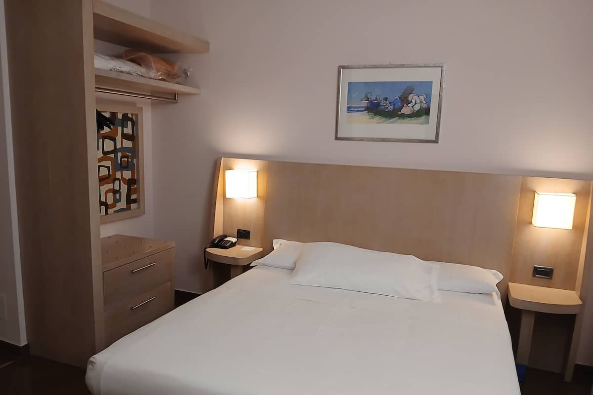 Een hotelkamer met een bed, nachtkastjes, een telefoon, een wandlamp en een lijst met een afbeelding.