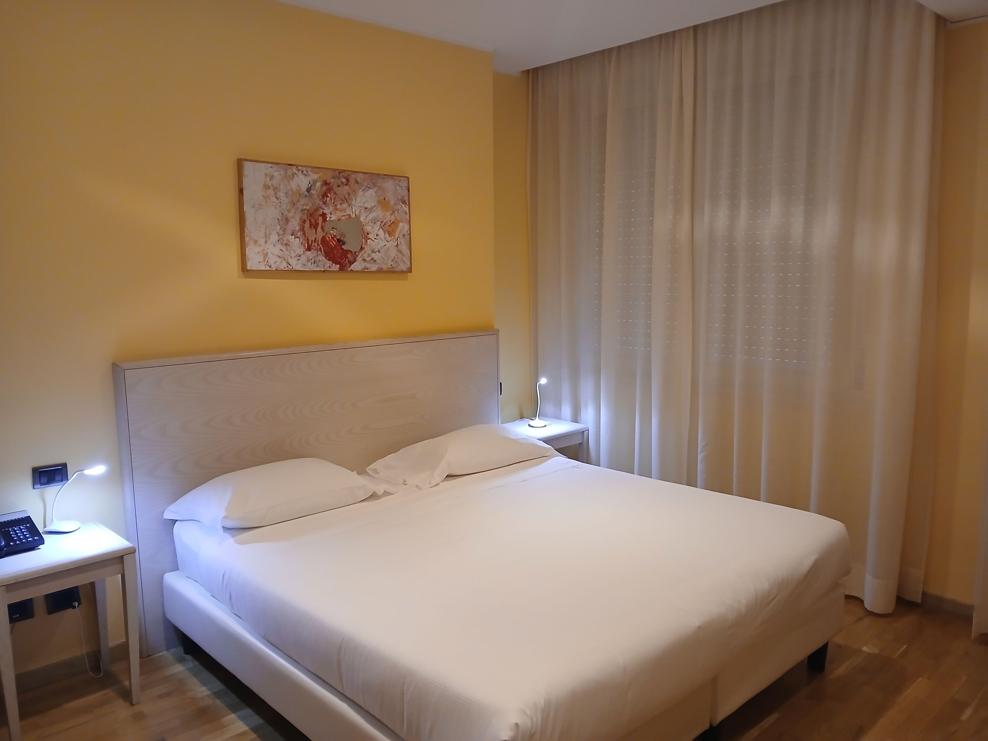 A neatly made bed with white linens, a bedside table with a lamp, and a wall-mounted artwork.