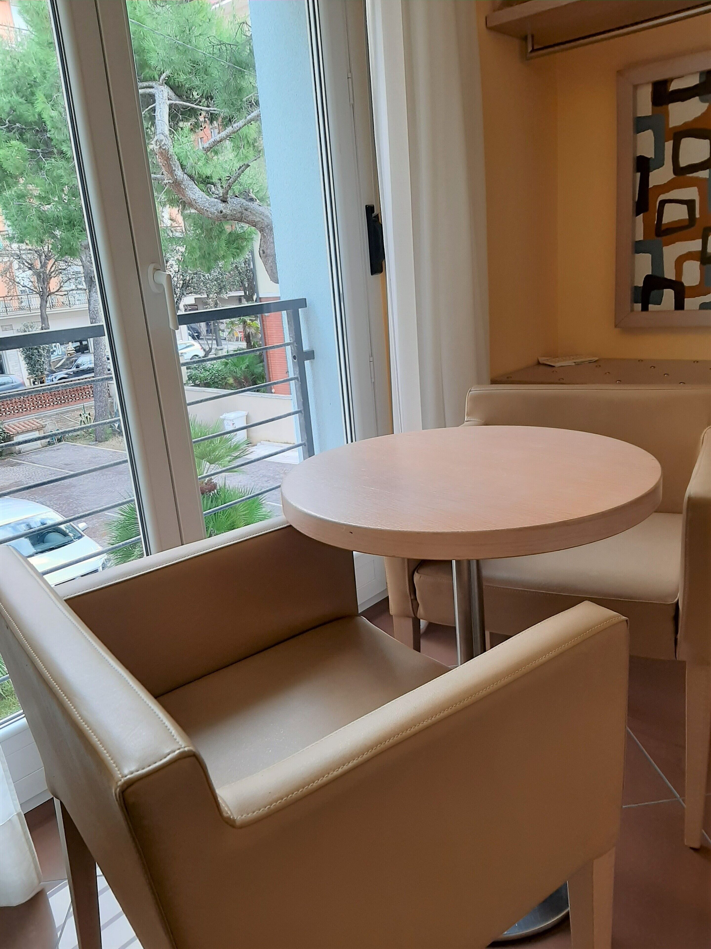 Una habitación con una mesa redonda y dos sillas color beige, una ventana que da a un balcón y árboles, y una obra de arte abstracta enmarcada en la pared.