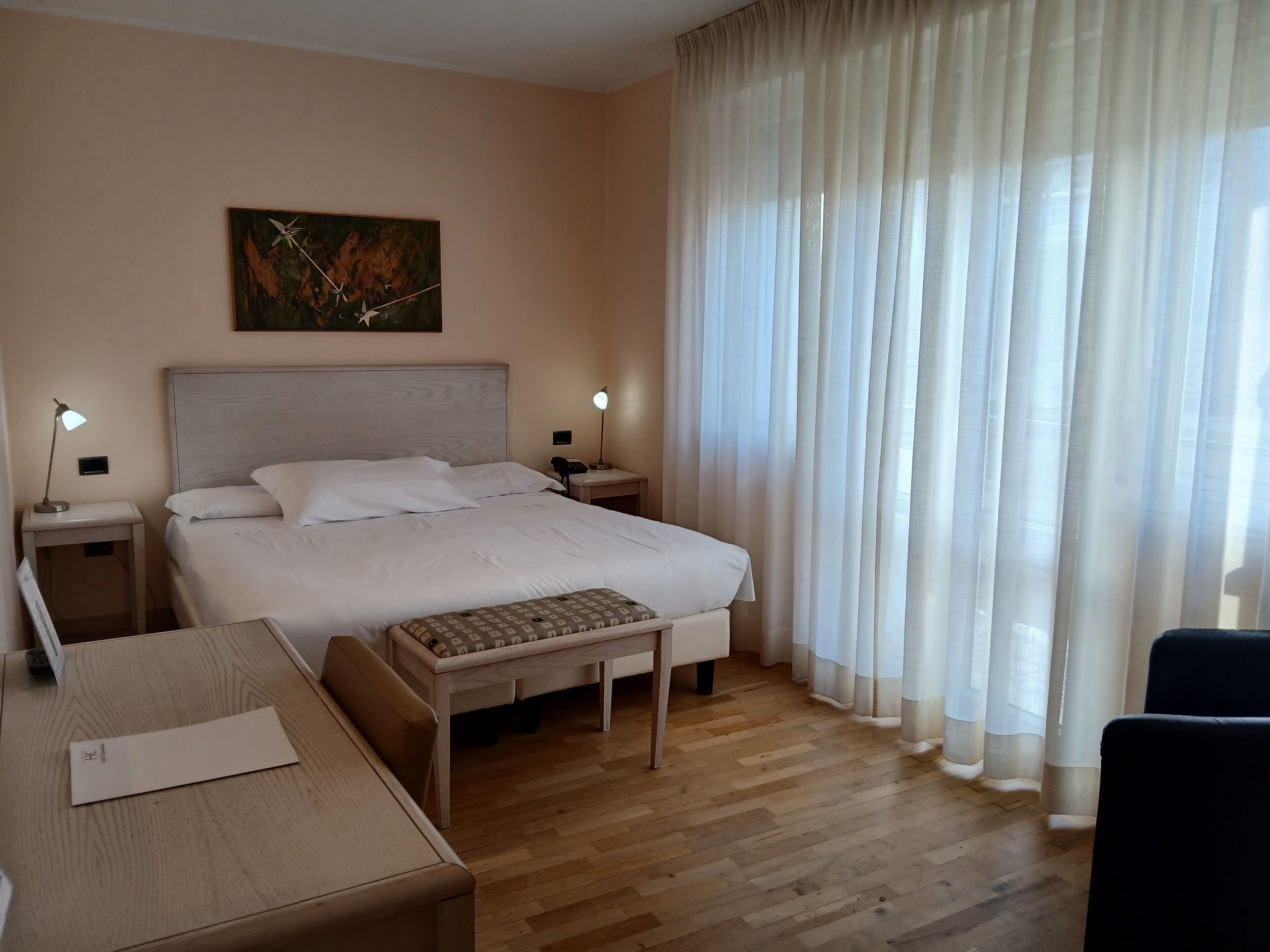 A hotel room with a bed, bedside tables, a desk, a chair, and a painting on the wall.