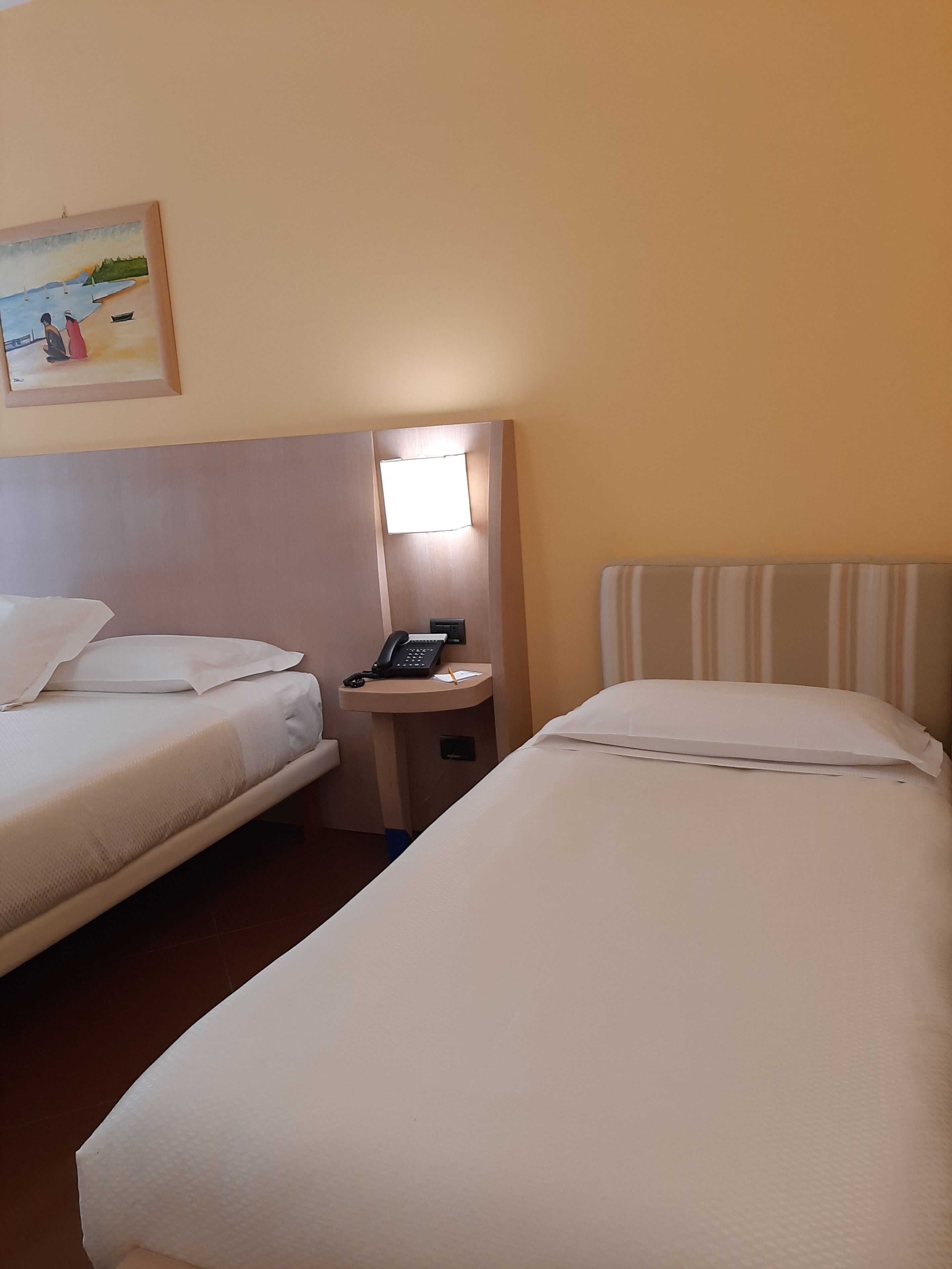 A hotel room with two single beds, a bedside table with a phone and lamp, and a painting on the wall.