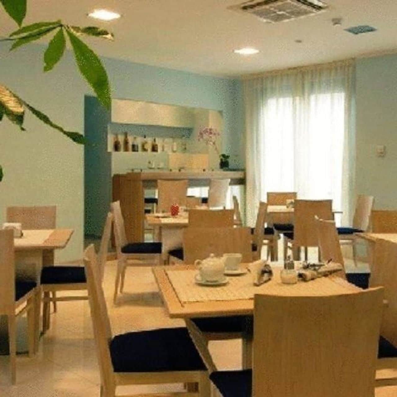 A dining area with wooden tables and chairs, a kitchen in the background, and a plant on the left.