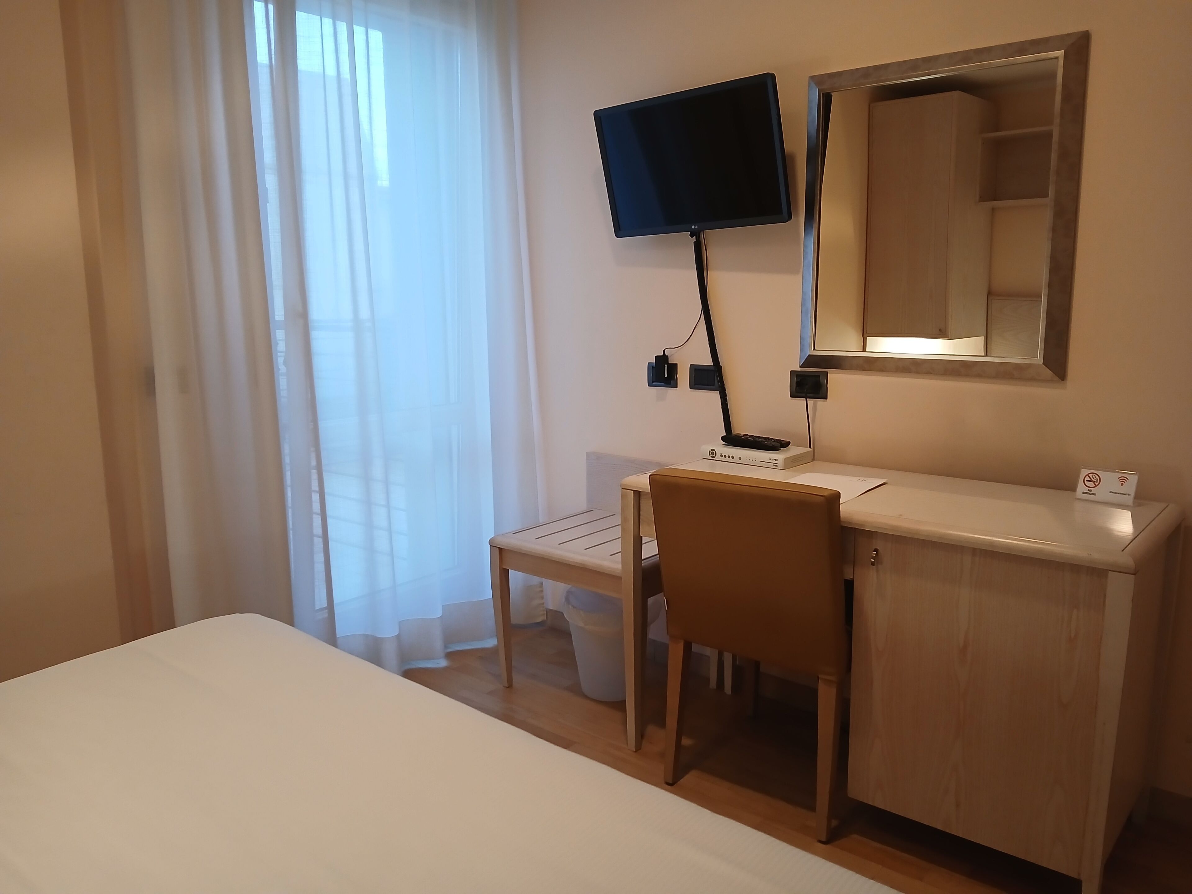 Habitación de hotel con cama, escritorio con computadora, televisor montado en la pared y ventana con cortinas translúcidas.