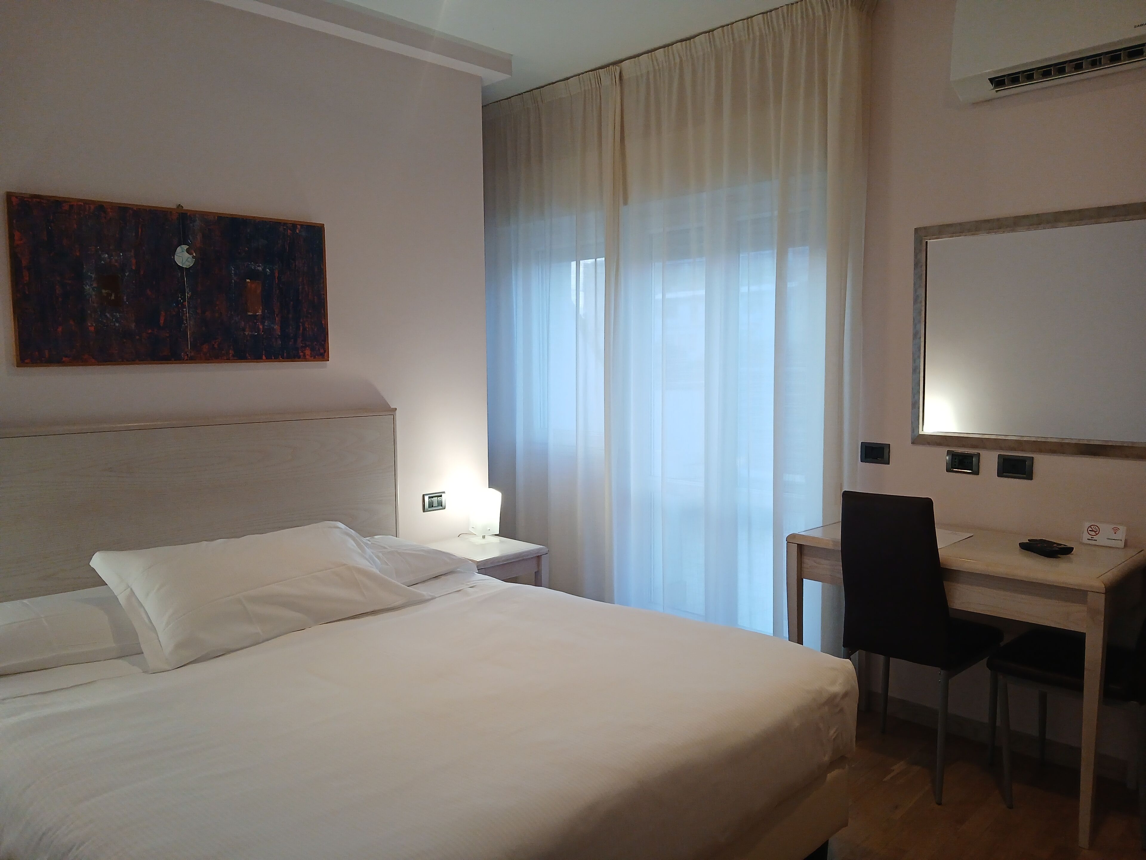Ein Hotelzimmer mit Bett, Schreibtisch, Stuhl, einem Bild an der Wand und einem Fenster mit Vorhängen.