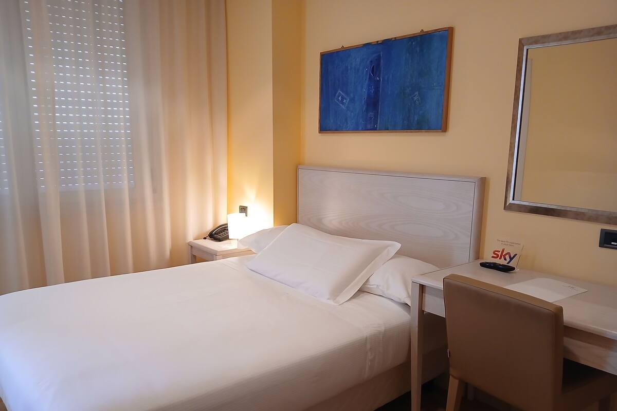 Een hotelkamer met een bed, nachtkastje, lamp, bureau, stoel en een schilderij aan de muur.