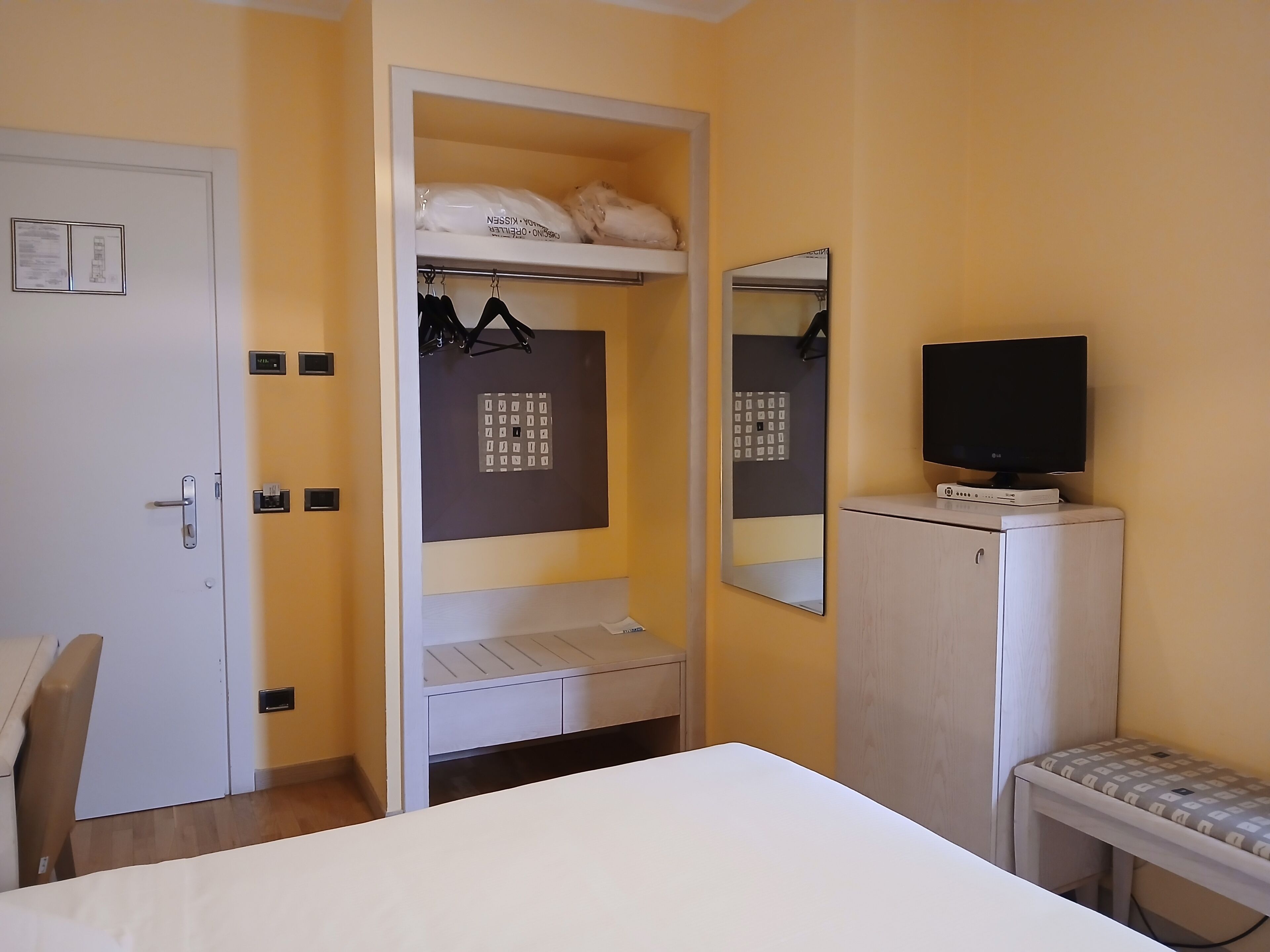 Une chambre d’hôtel équipée d’un lit, d’un bureau, d’une armoire, d’un miroir, d’une télévision et d’une étagère où sont suspendus des vêtements.
