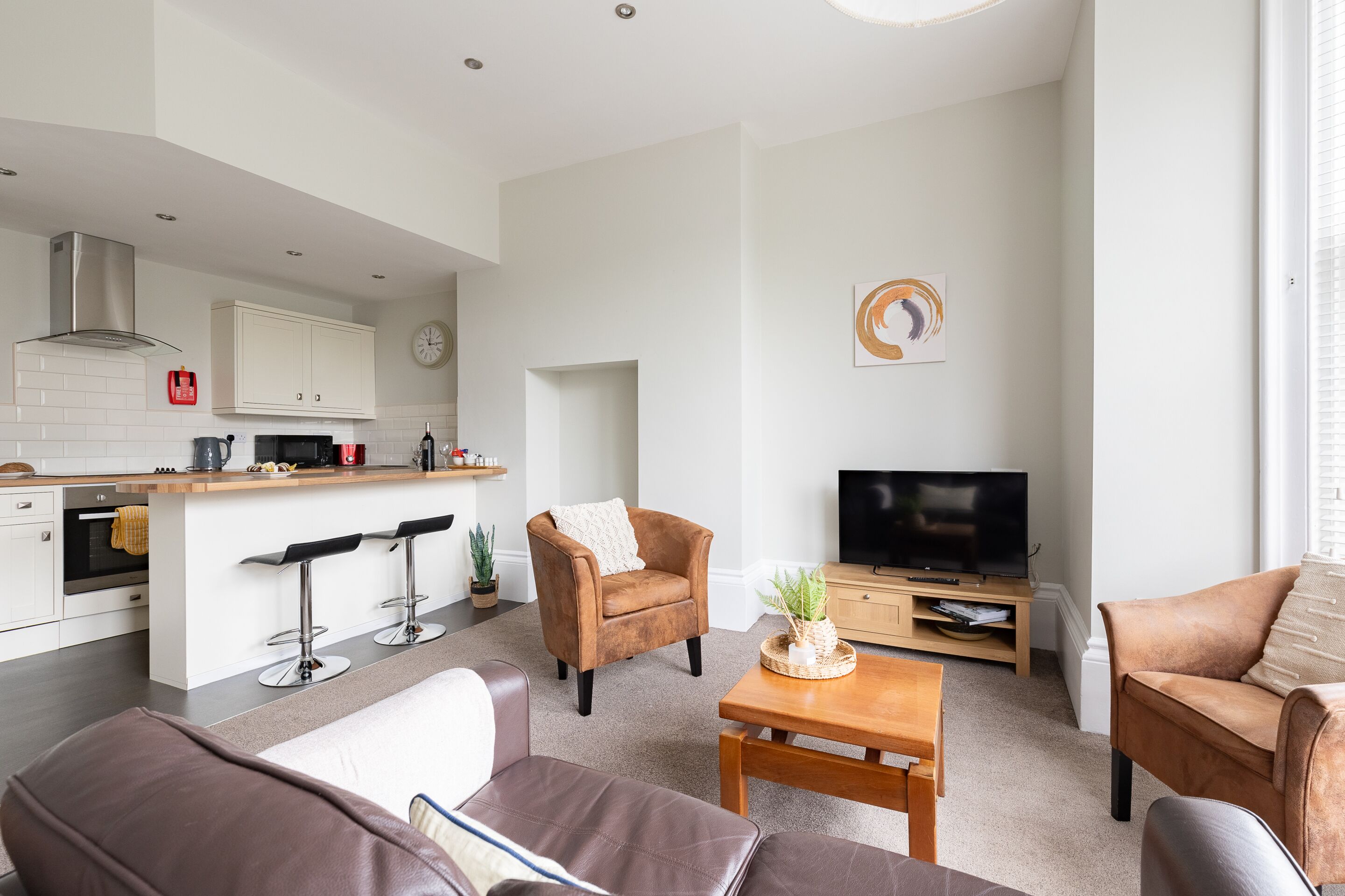 A modern kitchen with white cabinets, a built-in oven, and a stainless steel range hood. There is a dining area with a wooden table and two brown leather sofas. A television is mounted on the wall, and a plant is on the table.