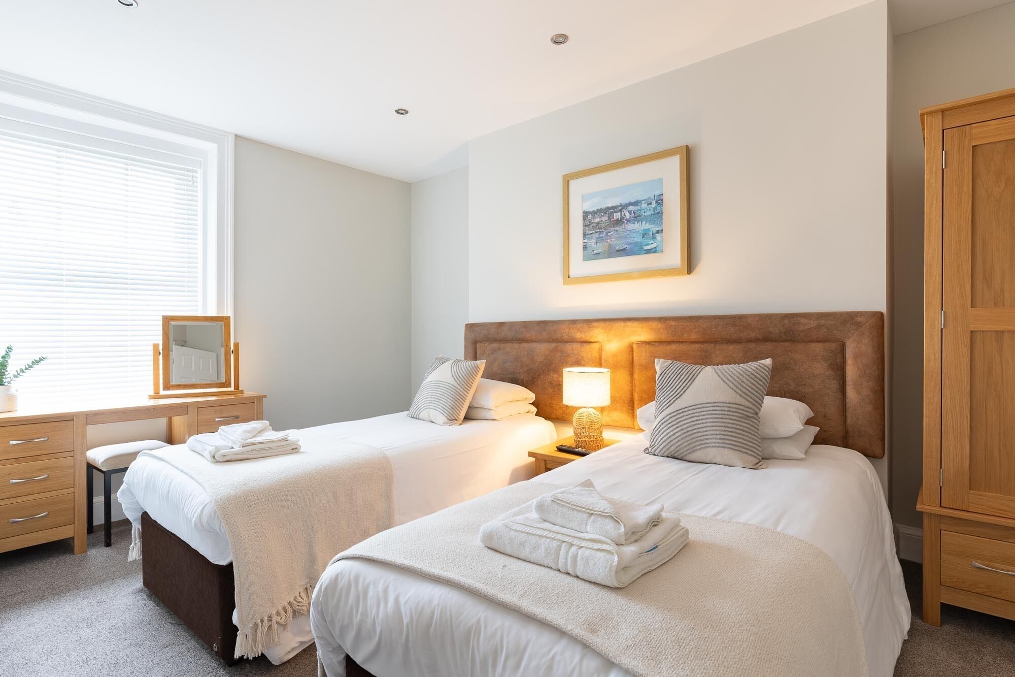 A hotel room with two beds, a wooden wardrobe, a desk with a mirror, and a framed picture on the wall.