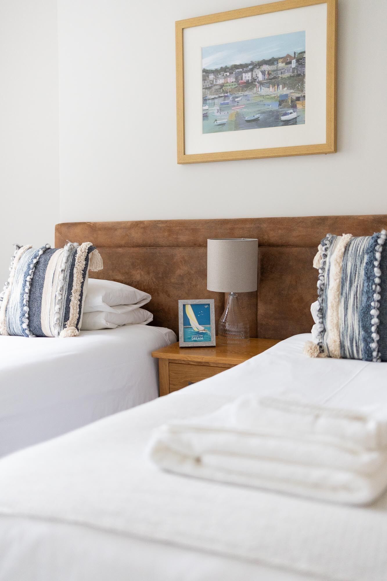 Une chambre d’hôtel avec deux lits, une tête de lit en bois, un tableau encadré au mur, une table de chevet avec une lampe et un petit tableau encadré posé sur la table.