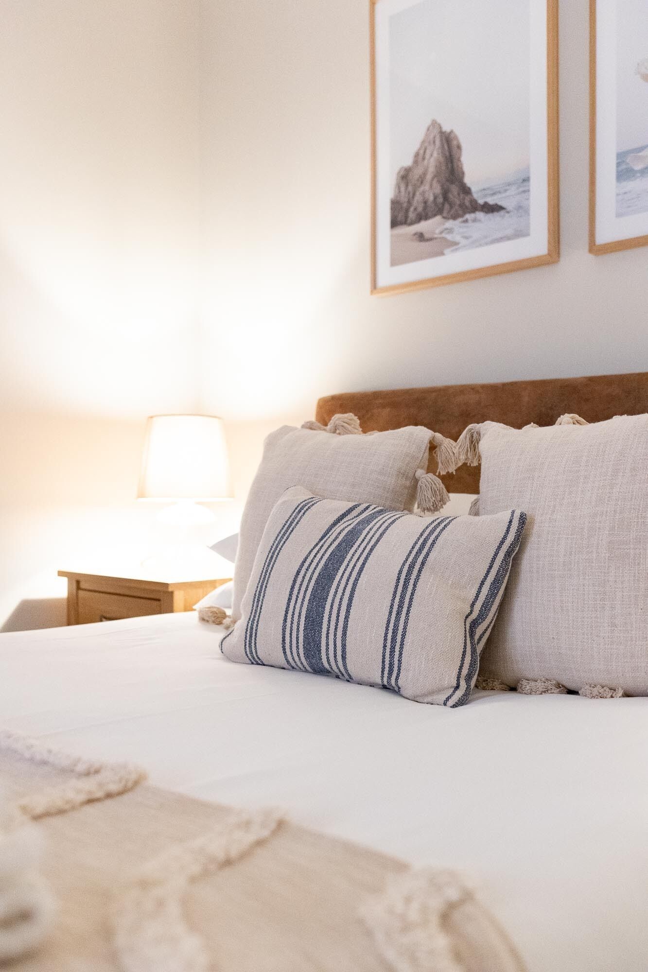 A neatly made bed with two pillows, a bedside table with a lamp, and two framed pictures on the wall.