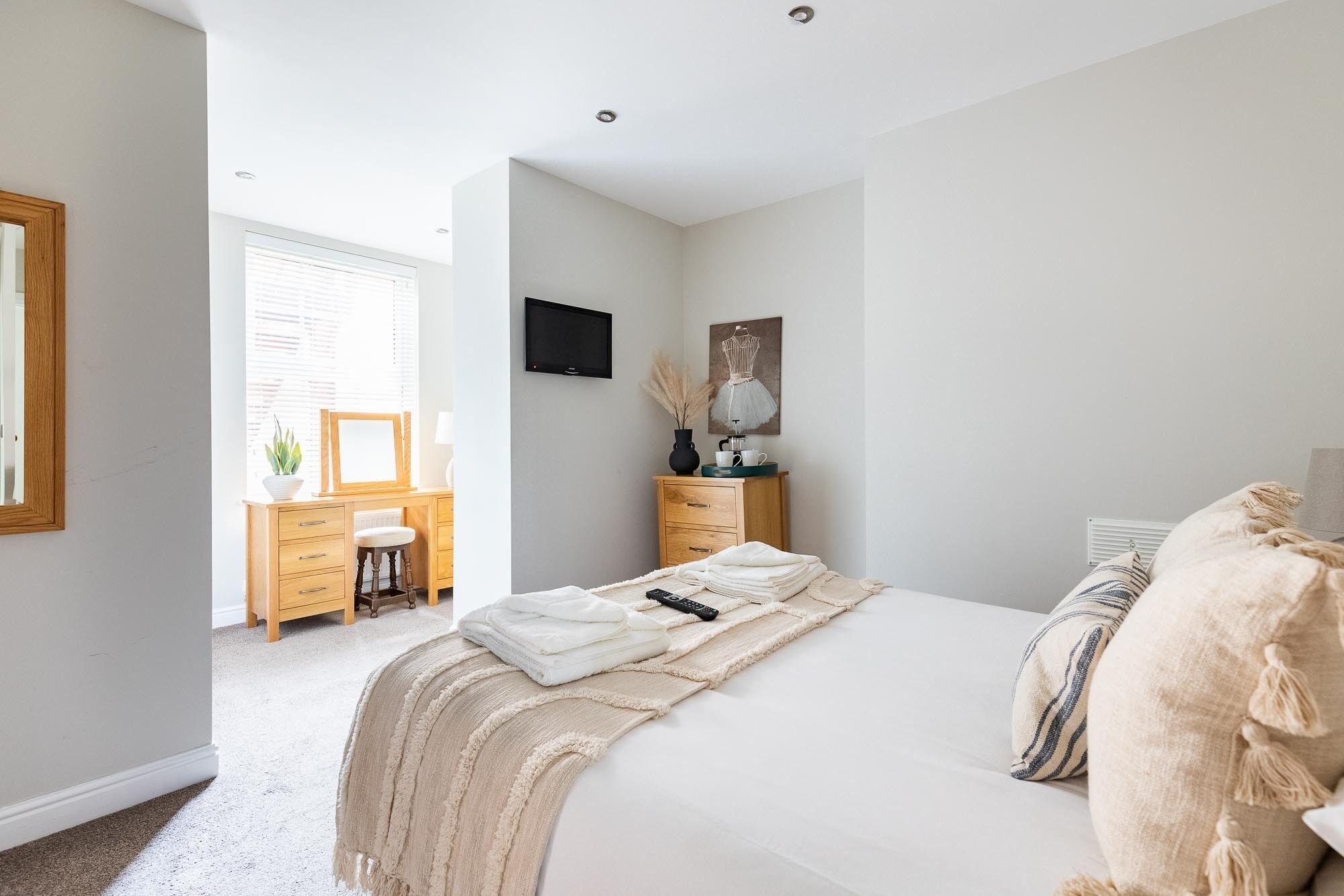 A bedroom with a bed, bedside table, and a dresser. There is a window with blinds, a TV mounted on the wall, and a painting above the dresser.