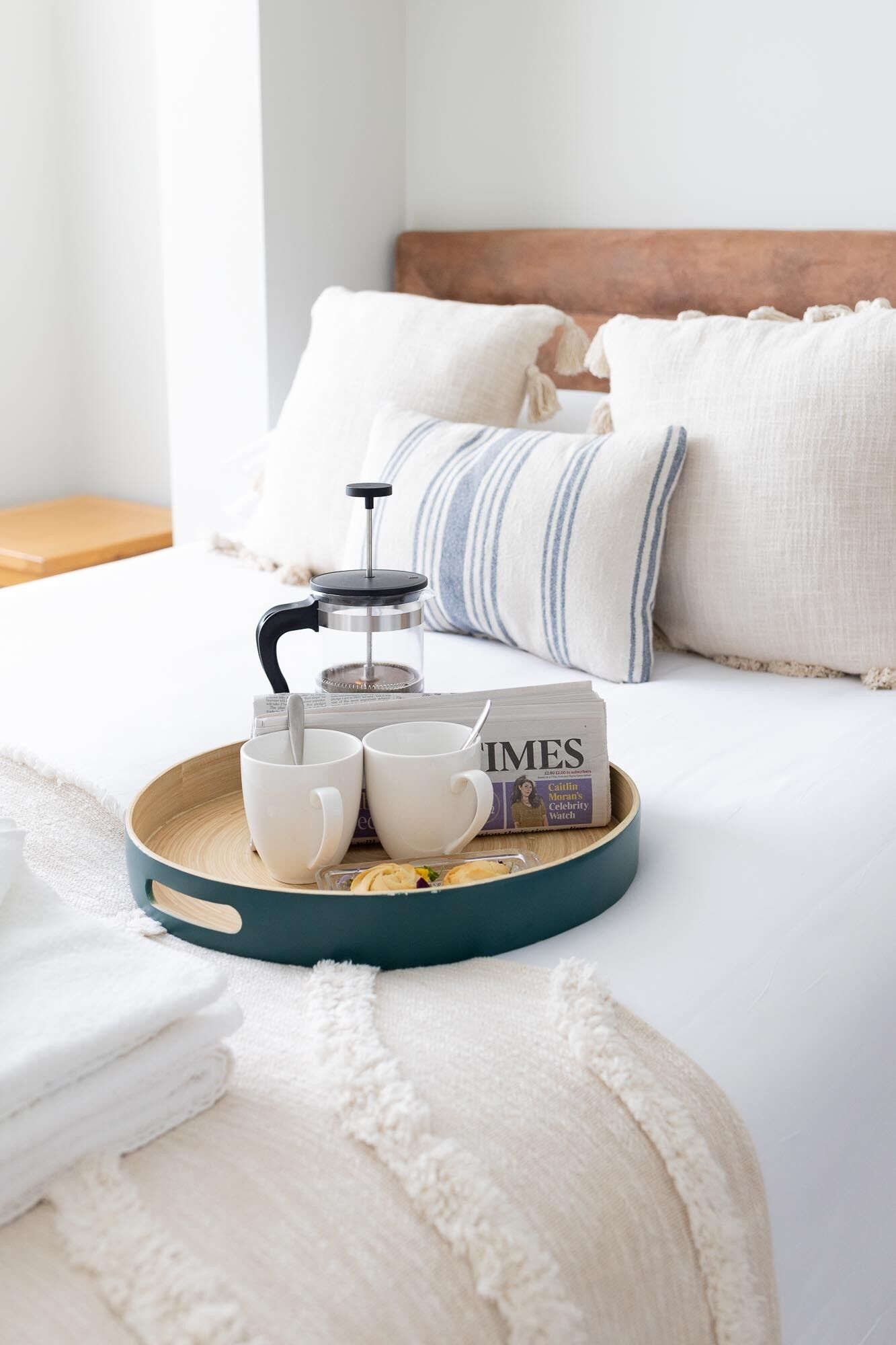 A neatly made bed with a wooden headboard, a tray with a coffee maker, two white cups, a Times newspaper, and a box of cookies.
