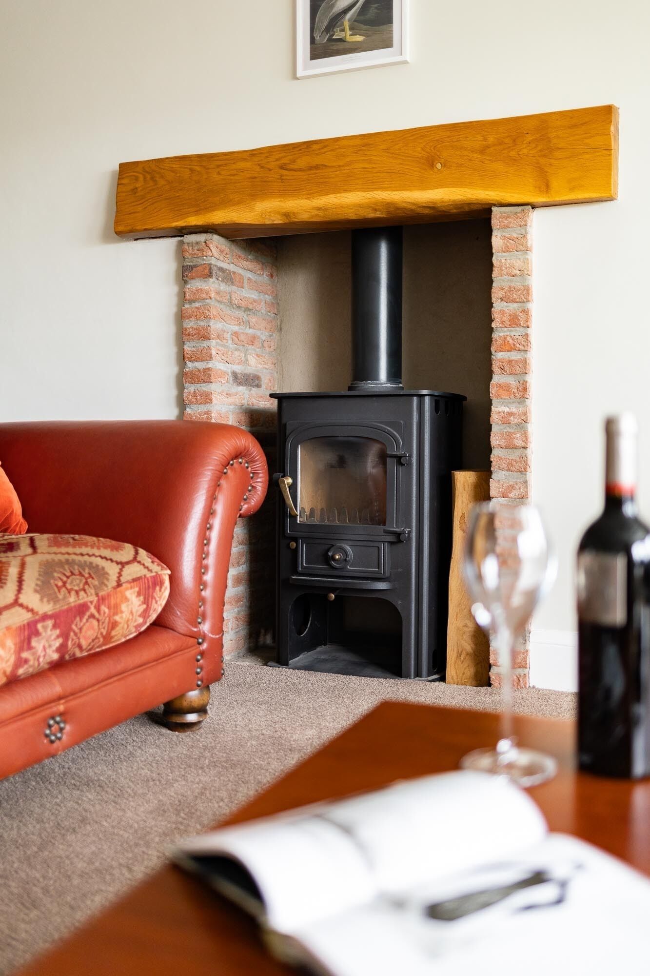 A cozy living room with a brick fireplace, a leather sofa, and a table with a wine glass and a book.