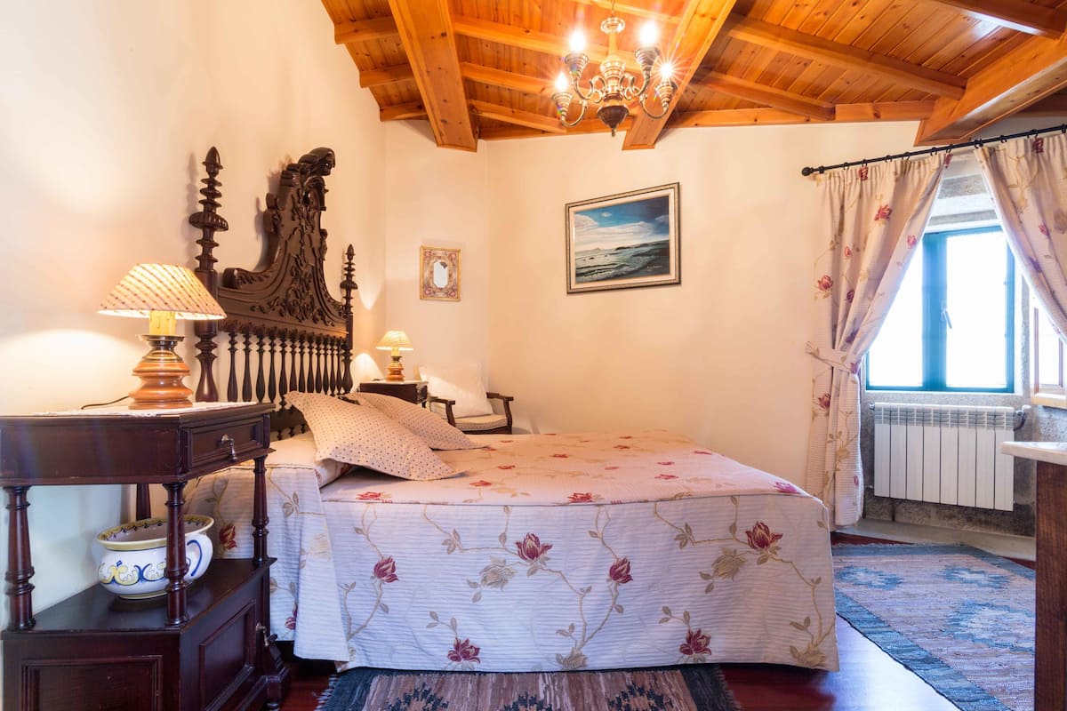 Un dormitorio con techo de madera, una cama con ropa de cama floral, una mesita de noche con lámpara y una ventana con cortinas.