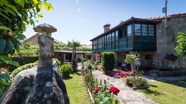 A stone building with a green balcony, surrounded by a well-maintained garden with various flowers and a stone statue. - Casa de Marcelo (Padron)