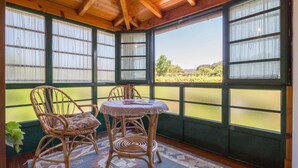 A room with a wooden ceiling, large windows with green frames, wicker chairs, a small table with a lace tablecloth, and a patterned rug. - Casa de Marcelo (Padron)