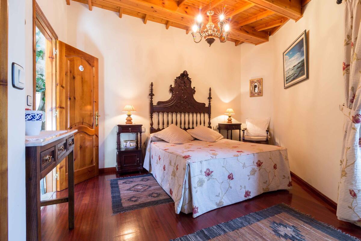 Un dormitorio con techo de madera, una cama grande con ropa de cama floral, dos lámparas de noche, una mesita de noche de madera y una puerta de madera.