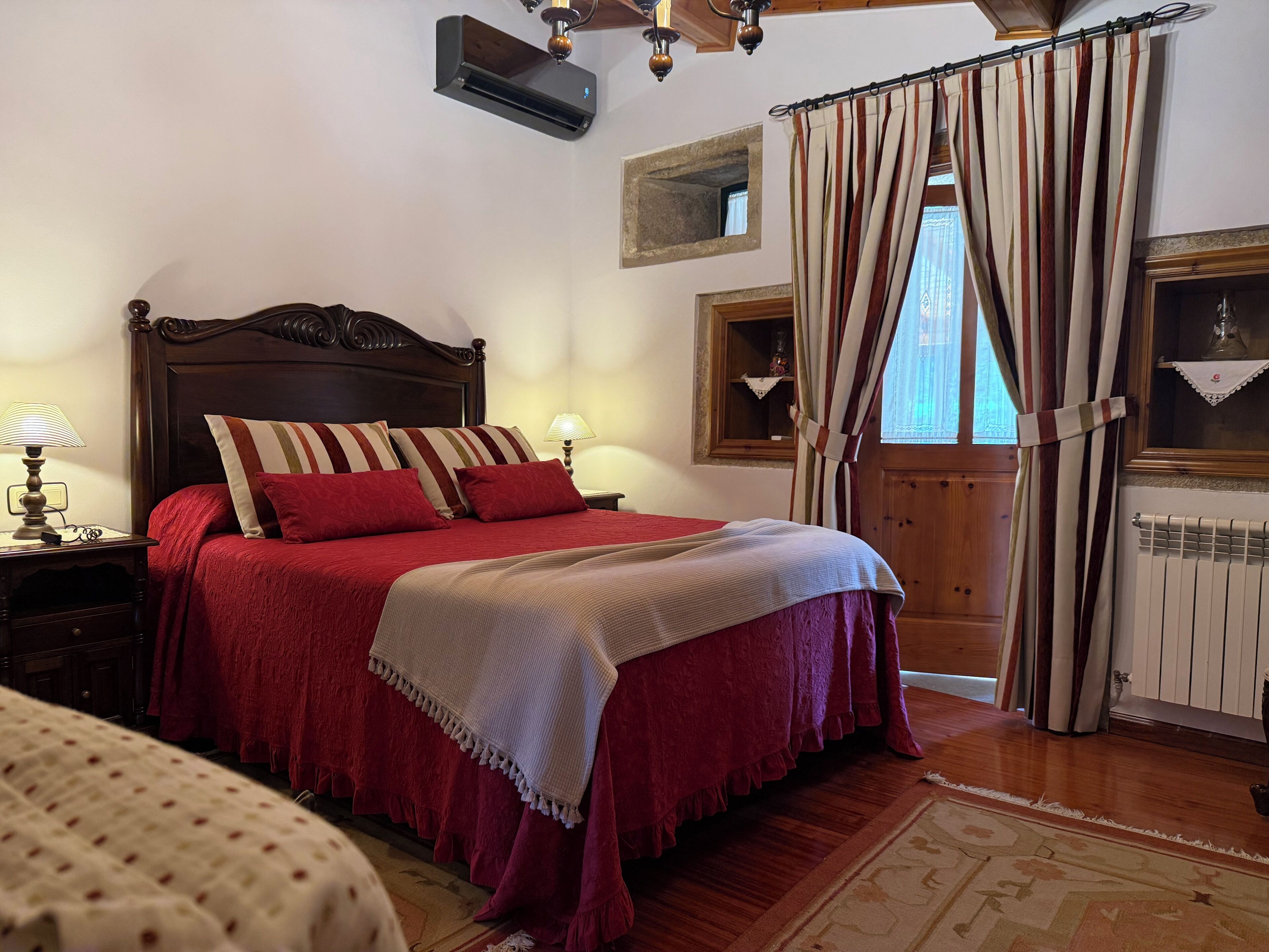 A bedroom with a wooden bed, red bedding, a nightstand with a lamp, a window with curtains, and a radiator.