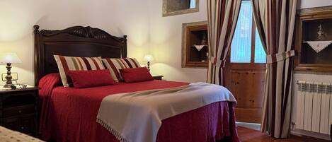 A bedroom with a wooden bed, red bedding, a nightstand with a lamp, a window with curtains, and a radiator.