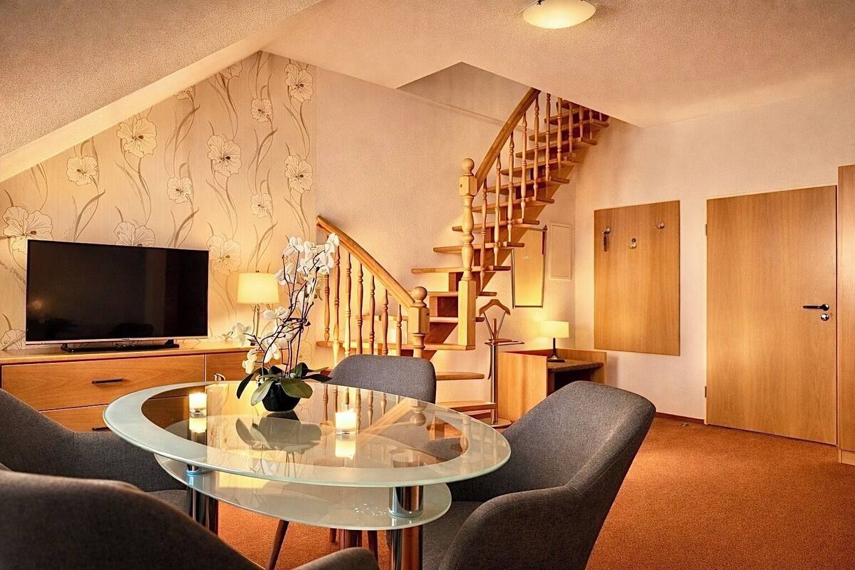 Un salon moderne avec une table à manger en verre, des canapés gris, un téléviseur, un escalier et des portes en bois.