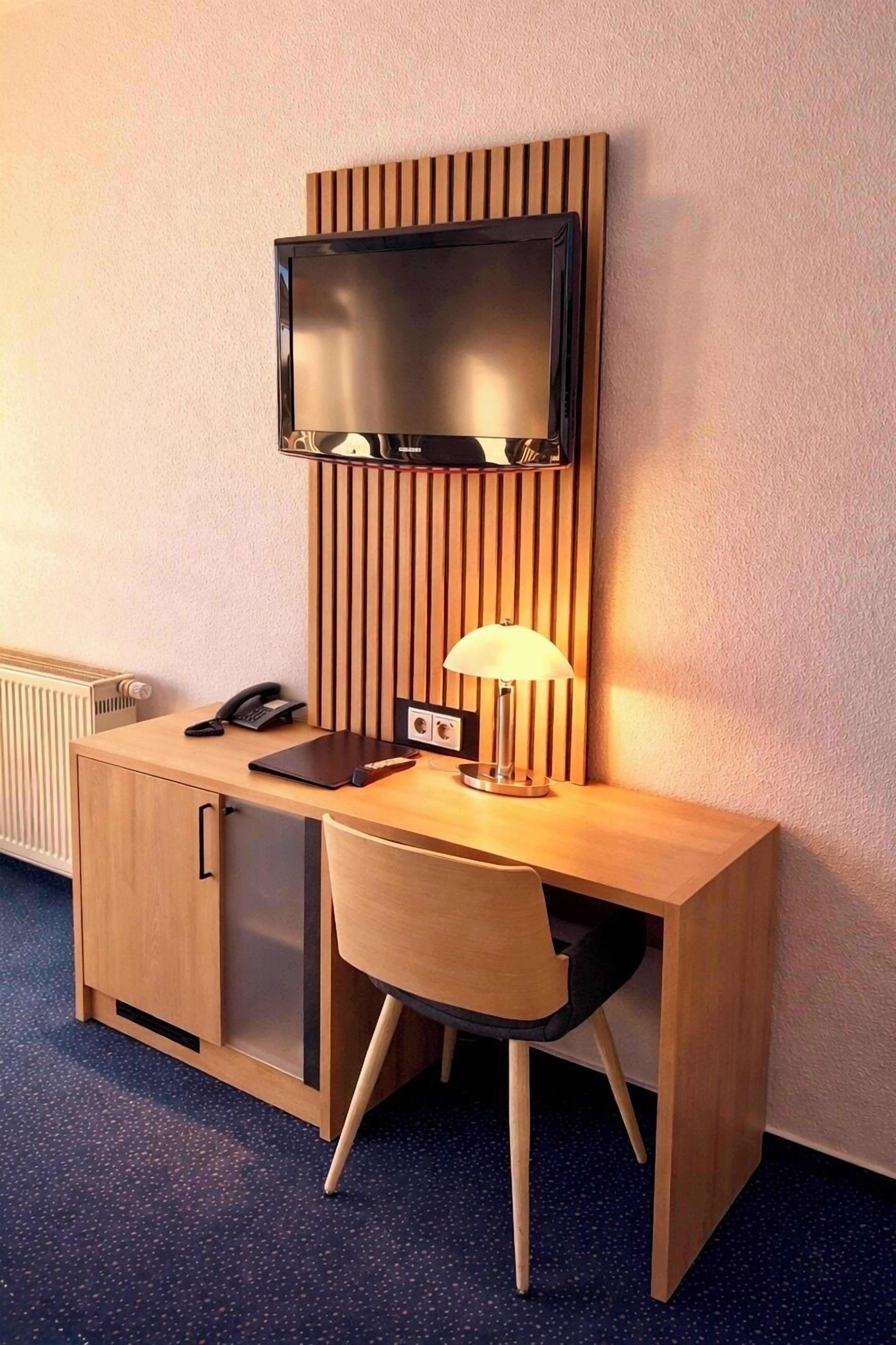 Habitación de hotel compacta con escritorio de madera, televisor montado en un panel ranurado, lámpara de escritorio y teléfono.