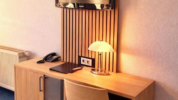 Habitación de hotel compacta con escritorio de madera, televisor montado en un panel ranurado, lámpara de escritorio y teléfono.
