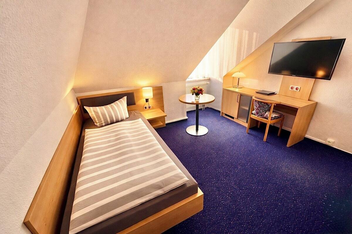 Une chambre d’hôtel de taille réduite, comprenant un lit, un bureau, une chaise, une petite table avec une lampe et un téléviseur fixé au mur.
