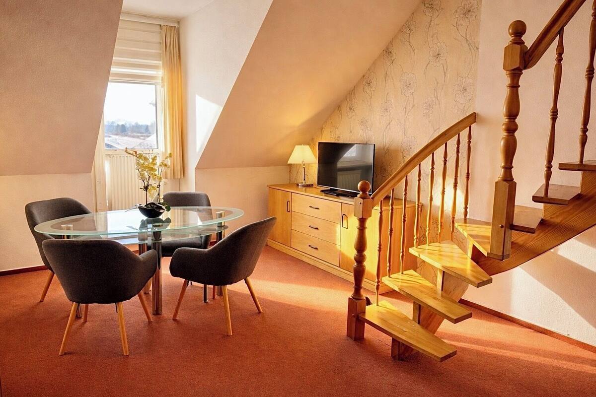 Un salon moderne avec une table à manger en verre, deux chaises, un meuble en bois, un téléviseur et un escalier en bois.