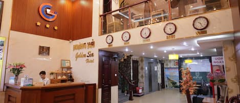 A hotel reception area with a wooden reception desk, a receptionist, and a staircase leading to an upper level.