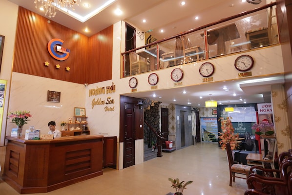 A hotel reception area with a wooden reception desk, a receptionist, and a staircase leading to an upper level.