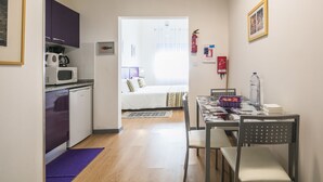 Habitación de hotel compacta con kitchenette, una cama con colcha estampada, una mesa de comedor con sillas y una pequeña alfombra.