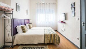 Un dormitorio ordenado con cama, mesita de noche, lámpara colgante, ventana con cortinas traslúcidas y una puerta.