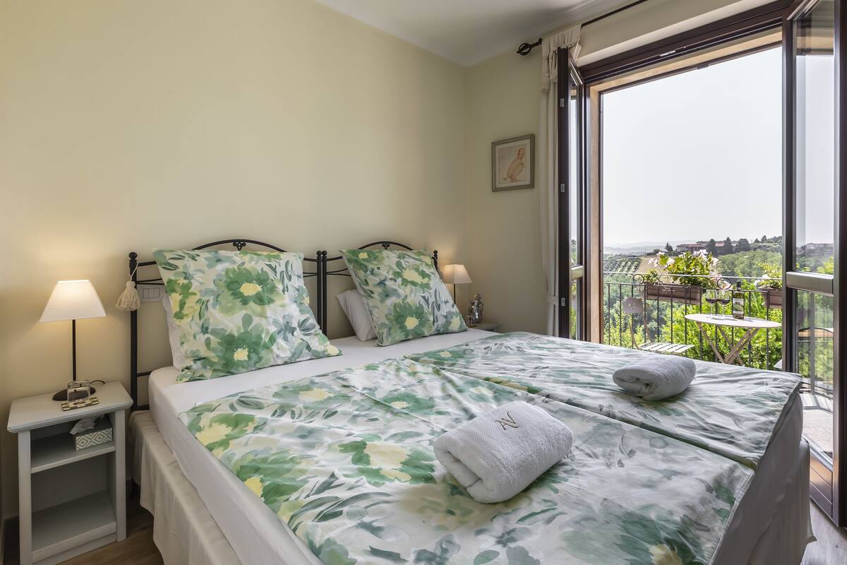 Un dormitorio con una cama grande, dos mesitas de noche con lámparas, una ventana con vistas a zonas verdes y edificios, y un balcón con una mesa y sillas.
