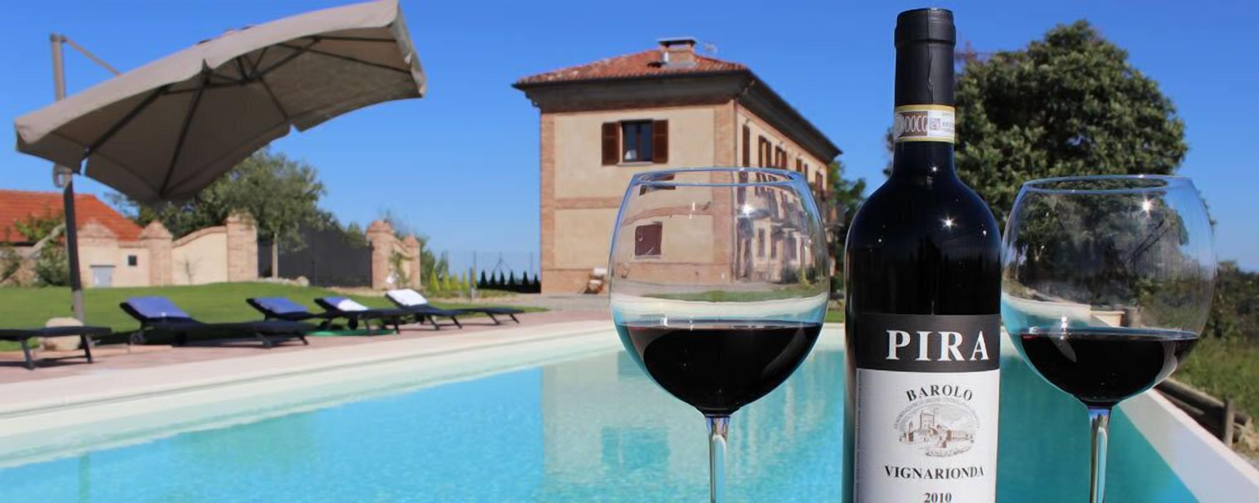 A poolside setting with a villa in the background, a bottle of Pira wine, and two glasses of wine.