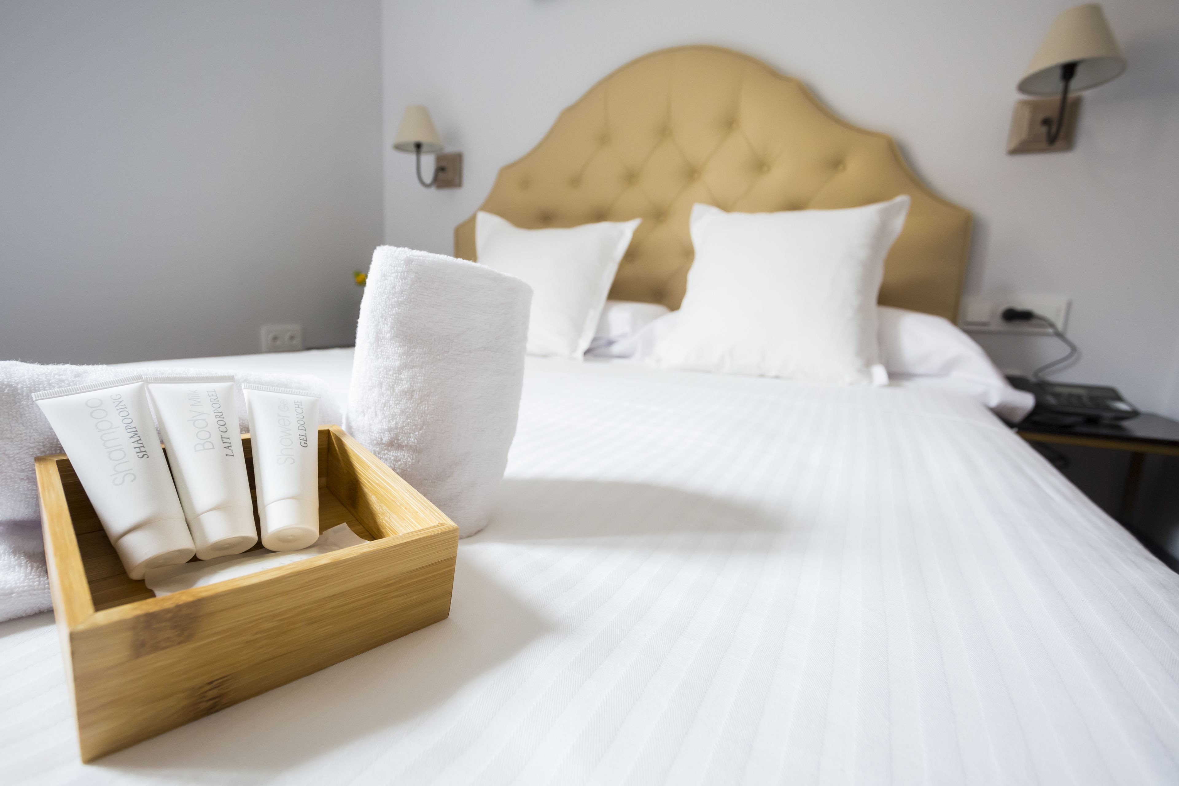 A hotel room with a neatly made bed, a wooden tray with toiletries, and a headboard with a light-colored fabric.