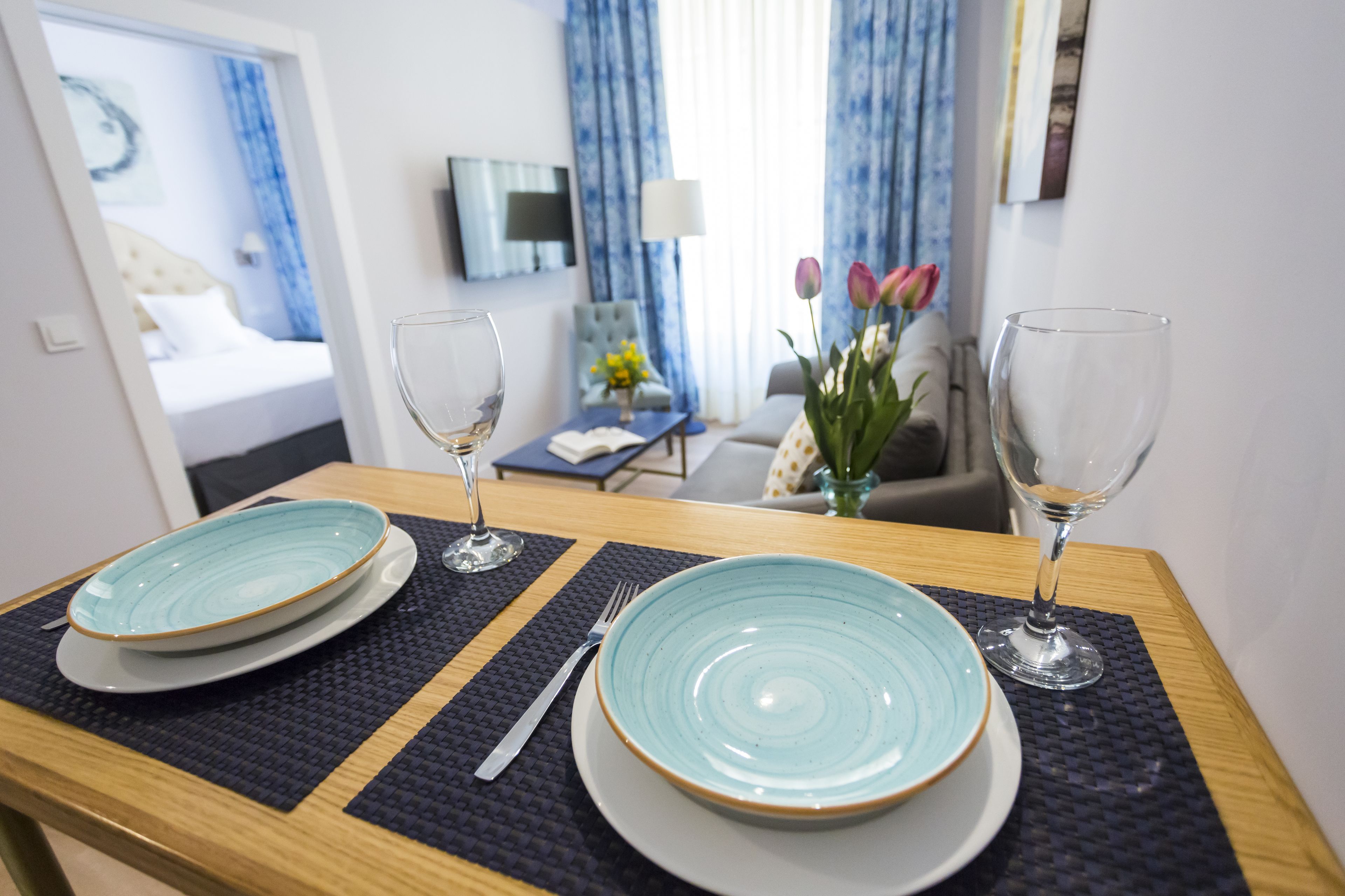 A hotel room with a dining table set with plates and wine glasses, a bed with white bedding, a TV mounted on the wall, and a vase with flowers on a side table.