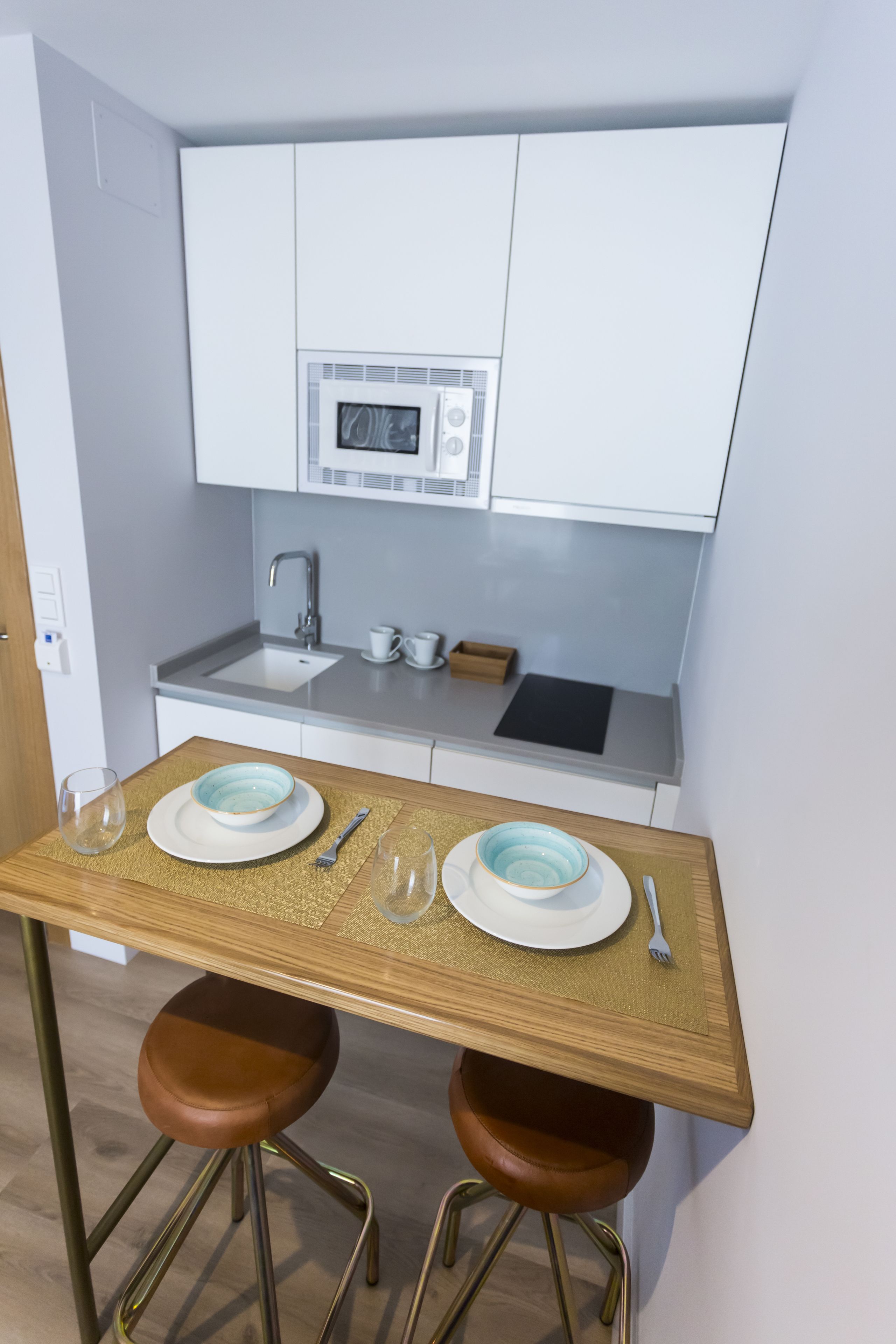 Cocina compacta con armarios blancos, microondas, lavaplatos y una pequeña encimera. Hay una mesa de madera con dos taburetes y algunos platos y utensilios.