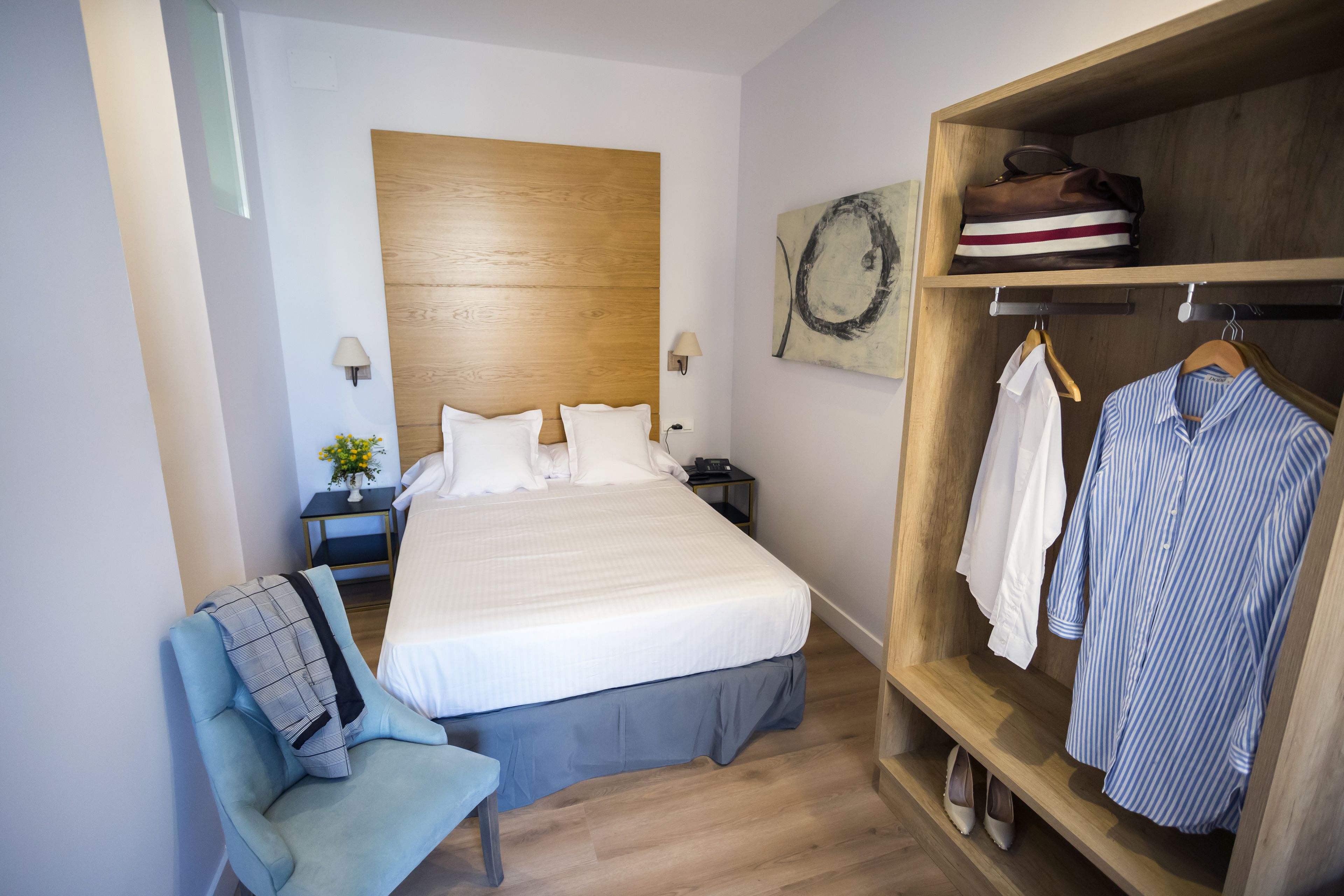 A bedroom with a bed, a wooden headboard, a bedside table with a lamp, a chair, and a wardrobe with hanging clothes.